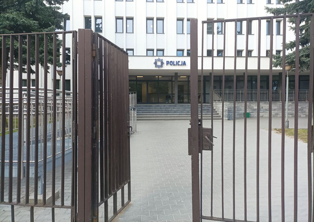 metalowe ogrodzenie z prętów oraz furtka prowadzące do budynku Komendy Miejskiej Policji w Zamościu , w tle budynek komendy z wejściem głównym przed którym są schody 