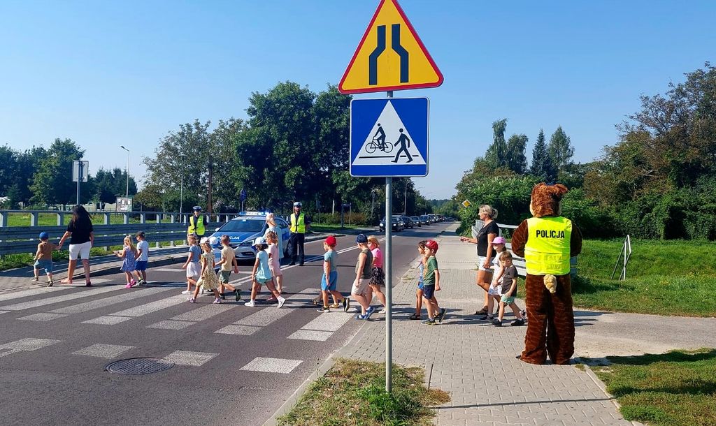 przed przejściem dla pieszych stoi policyjna maskotka, po pasach idą dzieci z opiekunkami 