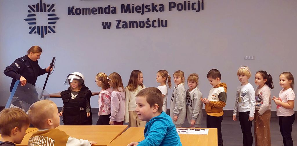 policjantka , dziecko w kasku ochronnym z tarczą policyjną oraz stojące w rzędzie inne dzieci