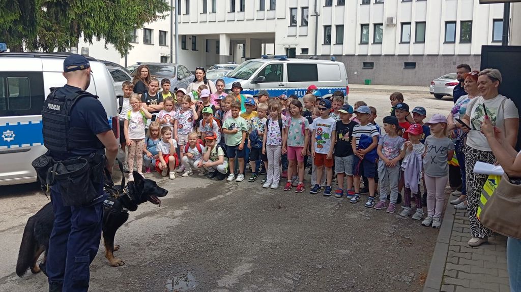 rozmowa policyjnego przewodnika z dziećmi, obok policjanta stoi pies , dalej w półkolu stoją dzieci 