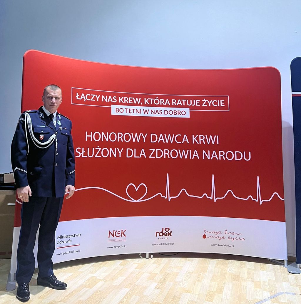 podinspektor Robert Oleszczuk w wyjściowym mundurze na tle ścianki medialnej Honorowi Dawcy krwi 