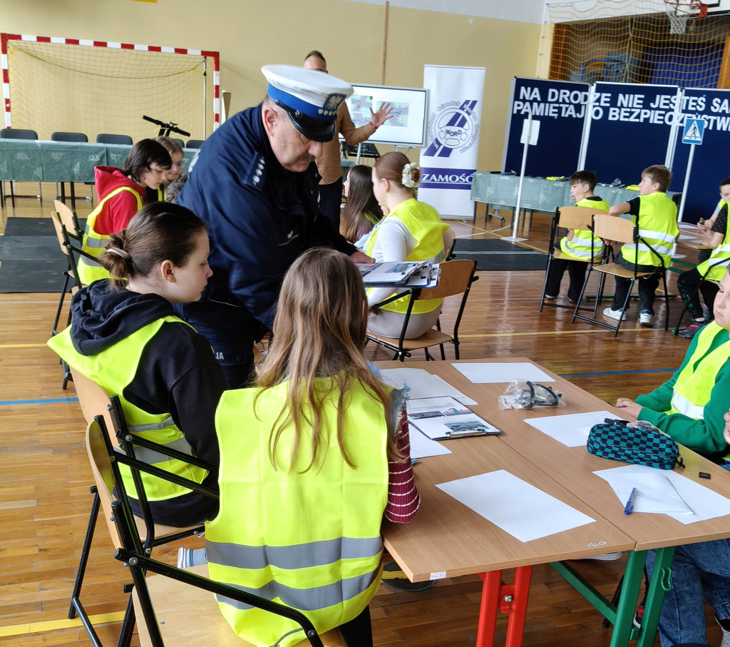 policjant obok stolika, przy którym na krzesłach siedzą uczniowie 