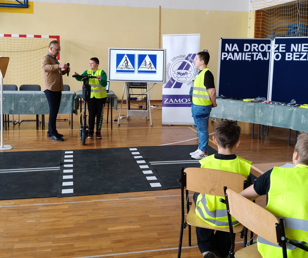 pracownik Wojewódzkiego Ośrodka Ruchu Drogowego obok dwóch chłopców na makiecie miasteczka ruchu drogowego, jeden z chłopców trzyma hulajnogę  