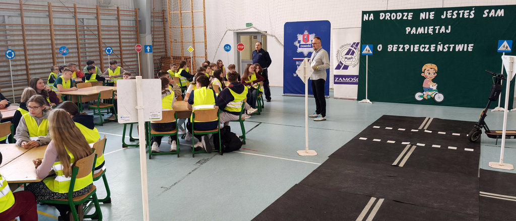 miasteczko ruchu drogowego w sali gimnastycznej szkoły, w tle stoi policjant i mężczyzna, przy stolikach siedzą uczniowie