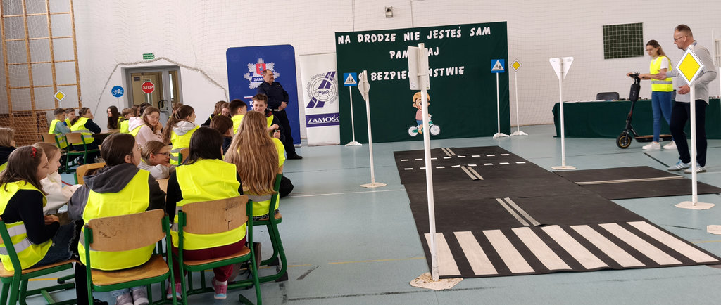 przy stolikach siedzą uczniowie mówią do nich policjant i mężczyzna, sala gimnastyczna szkoły, na której rozłożone jest miasteczko ruchu drogowego