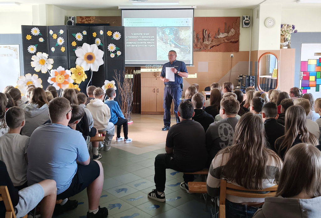 policjant stoi przed siedzącymi na krzesłach uczniami w czasie spotkania profilaktycznego 