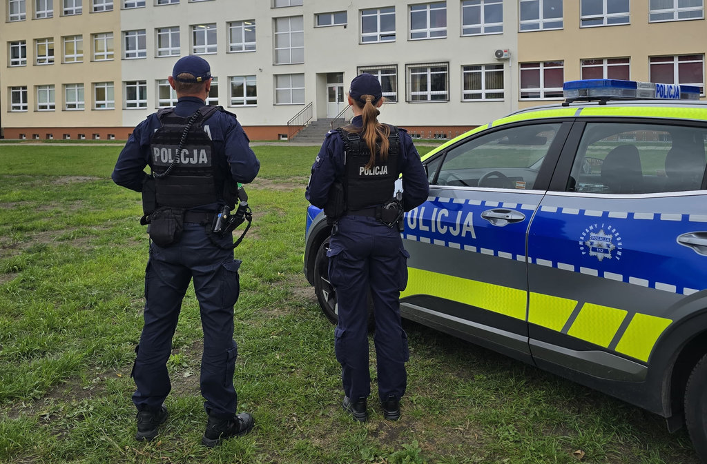 dwoje policjantów obok radiowozu na tle budynku szkoły 