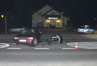 stojący na skrzyżowaniu samochód osobowy z uszkodzoną szybą czołową, obok leży motocykl, w tle policyjny radiowóz oraz budynek