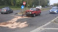 miejsce kolizji, uszkodzony czerwony samochód osobowy, w tle uszkodzony drugi pojazd osobowy w ciemnoszarym kolorze oraz stojący na ulicy policyjny radiowóz, na wysepce umiejscowionej na środku jezdni stoi znak drogowy zakaz wjazdu oraz nakaz jazdy po prawej stronie znaku