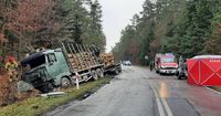 droga, miejsce tragicznego wypadku, w przydrożnym rowie stoi pojazd ciężarowy z uszkodzoną kabiną, obok strażak, po przeciwnej stronie jezdni rozłożony parawan straży pożarnej oraz pojazd strażacki, w tle policyjny radiowóz