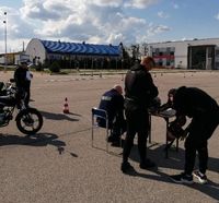 plac manewrowy z rozstawionym torem przeszkód gdzie odbywał się Ogólnopolski Młodzieżowy Turniej Motoryzacyjny, siedzący przy stoliku policjant, obok stoją dwaj uczestnicy Turnieju, w tle motocykl oraz policjant