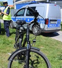 stojący na trawie rower, w tle policyjny radiowóz i umundurowana policjantka