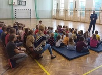 Zdjęcie w czasie prelekcji z uczniami w sali gimnastycznej. Dzieci siedzą na materacach, przed nimi stoi policjant.