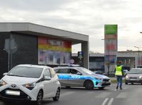 Miejsce potrącenia pieszej. W ulicy poprzecznej przed pasami zaparkowany radiowóz. Na poboczu drogi stoi osobowa toyota. Jezdnią idzie nieumundurowany policjant w kamizelce odblaskowej.