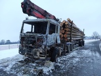 Stojąca na poboczu ciężarówka z naczepą na której załadowane są bele drewna. Kabina pojazdu jest spalona.