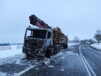 Stojąca na poboczu ciężarówka z naczepą na której załadowane są bele drewna. Kabina pojazdu jest spalona.