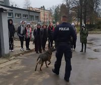 Policjant z psem służbowym oraz młodzież szkolna w czasie spotkania profilaktycznego.