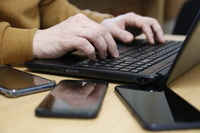 ręce trzymane na klawiaturze laptopa, obok leżą trzy telefony komórkowe