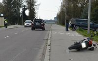 Na drodze stoi osobowy nissan. Przed nim na chodniku leży motocykl. Po drugiej stronie jezdni stoi umundurowany policjant.