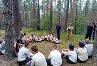 Las, siedzący harcerze i stający obok policjant i strażacy podczas spotkania profilaktycznego.