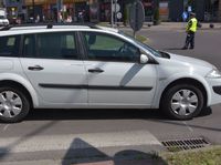 Na przejściu dla pieszych bokiem stoi osobowy renault. W tle na rondzie stoi policjant kierujący ruchem.