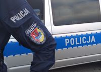 rękaw policyjnego munduru z naszywką logiem Komendy Miejskiej Policji w Zamościu, w tle radiowóz z bocznym napisem Policja