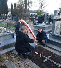 dwaj policjanci w ubraniach cywilnych podczas porządkowania grobu na krzyż zawieszają biało - czerwoną szarfę