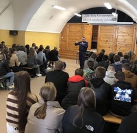 Stojący przed siedzącą na ławkach młodzieżą policjant w czasie spotkania profilaktycznego.