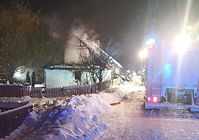 Dom drewniany i wydobywający się z jego wnętrza dym, obok stoją dwaj strażacy. Na zaśnieżonej ulicy stoi wóz strażacki.