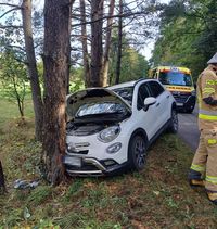 osobowy Fiat, który uderzył w drzewo. W tle karetka pogotowia. Obok fiata stoi strażak