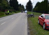 Na poboczu drogi stoi osobowa skoda, we wjeździe na posesję motorower. Na ulicy stoi policjant a w oddali radiowóz.