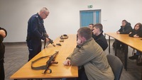 policjant pokazuje siedzącym na krzesłach za stołem słuchaczom jednostki broni