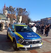 Na pierwszym planie policyjny radiowóz zaparkowany na płycie placu. Za radiowozem na placu stoją Komendant Miejski Policji w Zamościu inspektor Andrzej Mioduna, Komendant Komisariatu Policji w Szczebrzeszynie nadkomisarza Robert Oleszczuk, ksiądz oraz przedstawiciele samorządów.