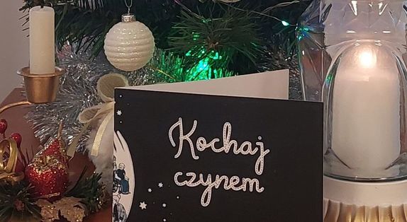 napis na kartce kochaj czynem, w tle świąteczne ozdoby i światło pokoju