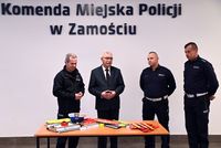 Starosta Zamojski Stanisław Grześko, Komendant Miejski Policji w Zamościu inspektor Andrzej Mioduna oraz dwóch Naczelników Wydziału Ruchu Drogowego stoją obok stolika, na którym leżą odblaskowe odblaski oraz czapka policjantów ruchu drogowego