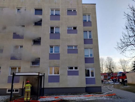 Blok mieszkalny, z okien klatki schodowej wydobywają się kłęby dymu. Przed drzwiami klatki schodowej stoi strażak. Obok bloku stoją trzy strażackie wozy bojowe.