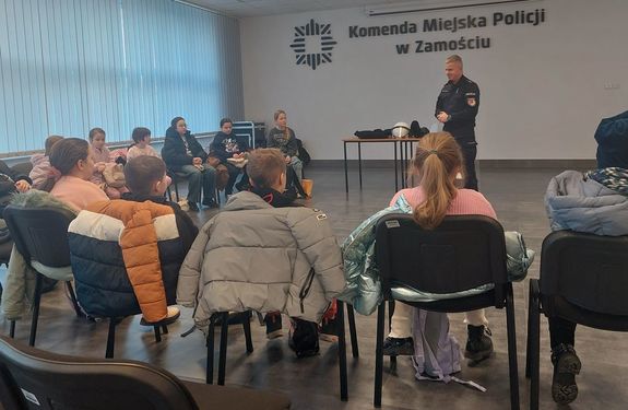 policjant stoi przed dziećmi siedzącymi na krzesłach w czasie spotkania profilaktycznego