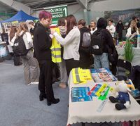 stoisko promocyjne Komendy Miejskiej Policji w Zamościu w czasie targów edukacyjnych, młodzież przymierza kamizelki odblaskowe