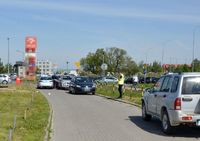 droga i zaparkowane na jej poboczu samochody, w tle policjanci wykonujący oględziny miejsca potracenia osoby pieszej