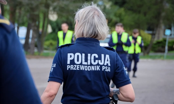 odwrócona tyłem policjantka z napisem na koszulce Policja przewodnik pas, w tle inni policjanci