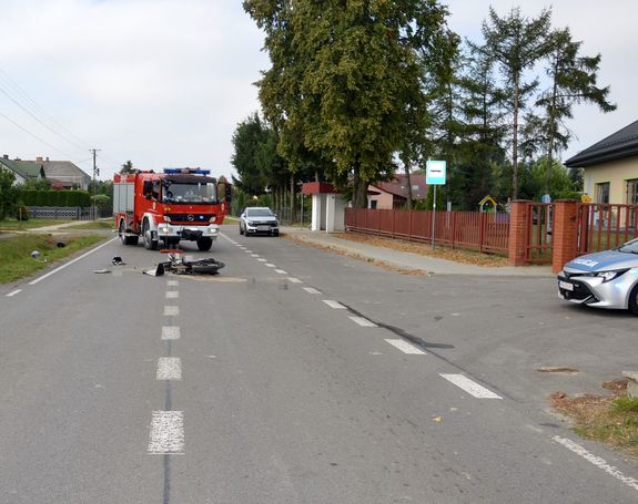 droga, miejsce wypadku, na jezdni leży motorower, w bocznej uliczce stoi radiowóz, za motorowerem stoi wóz strażacki oraz osobowa Dacia