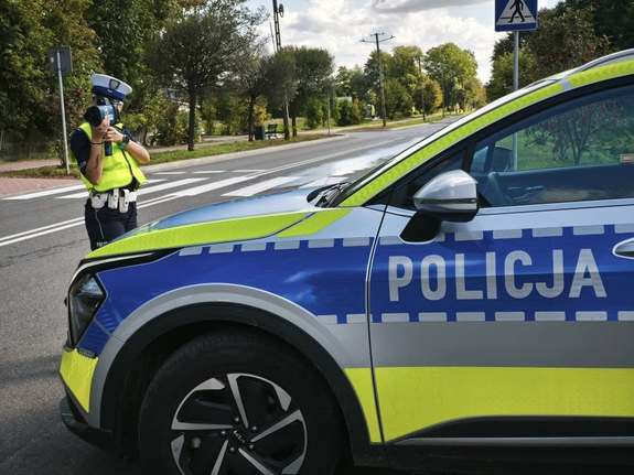 umundurowana policjantka w kamizelce odblaskowej dokonująca pomiaru prędkości przy pomocy miernika prędkości, przed nią widać oznakowany radiowóz policyjny