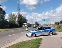policjant z pobocza drogi urządzeniem mierzy prędkość, obok niego stoi radiowóz