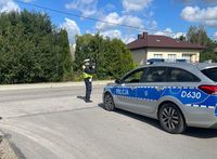 Policjant ruchu drogowego stoi na poboczu mierząc prędkość jazdy kierujących pojazdami. Obok policjanta zaparkowany jest radiowóz.