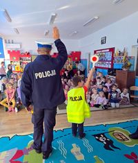 policjant i dziecko w kamizelce odblaskowej stoją przed siedzącą na dywanie grupą dzieci w czasie spotkania profilaktycznego w przedszkolu