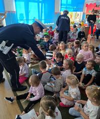 policjanci rozdają siedzącym na dywanie dzieciom książeczki