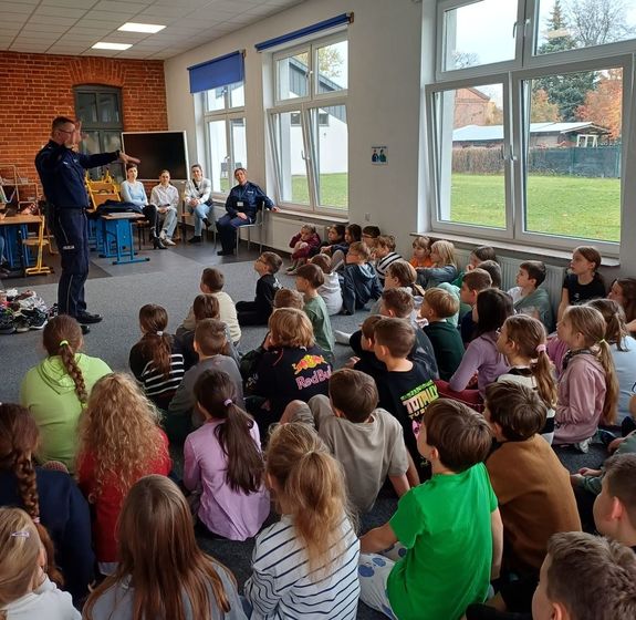 policjant rozmawiający z siedzącymi na dywanie i krzesełkach dziećmi, w czasie spotkania profilaktycznego