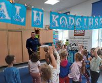 policjant rozmawiający z siedzącymi na dywanie i krzesełkach dziećmi, w czasie spotkania profilaktycznego