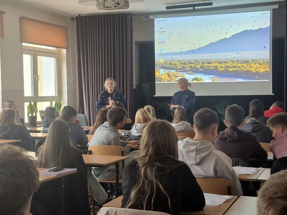 dwie policjantki w czasie spotkania profilaktycznego w szkole rozmawiają z siedzącą na krzesłach młodzieżą