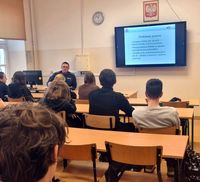 lekcja w klasie o profilu mundurowym, policjant siedzi za biurkiem a młodzież na krzesłach przy ławkach